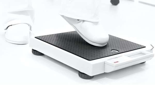 Primer plano de un pie tocando suavemente la base de la balanza SECA 876 para activarla automáticamente. Diseño de encendido sin botones para mayor comodidad en hospitales y clínicas.